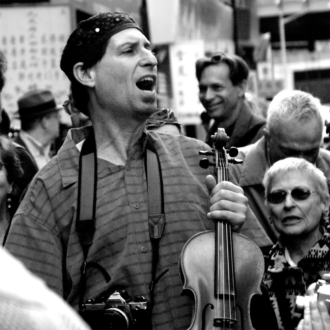 Man with a camera hanging around his neck and a violin in his hand singing amongst a crowd outside