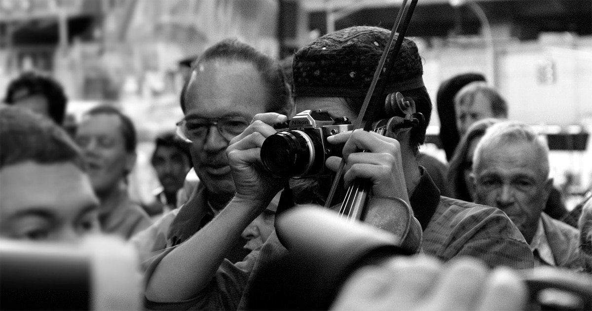 Man taking a picture with a big camera obscuring his face, standing in a crowd (black and white)