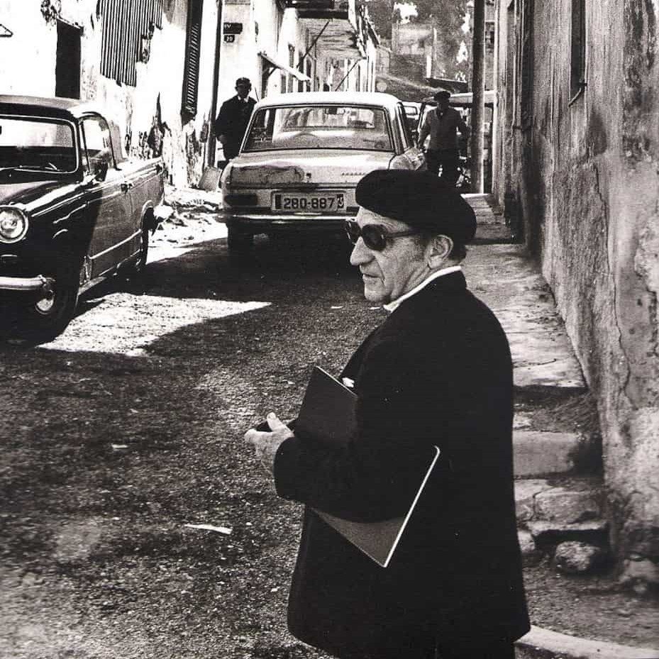 An old black and white photograph of Nahum Gutman with sunglasses and a beret, with a drawing pad under his arm. In the background a narrow street with cars and two men.